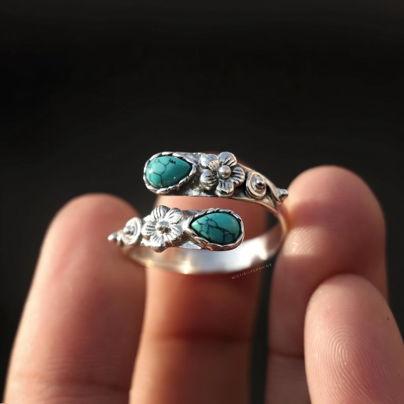 May include: A silver-toned ring with a floral design and two turquoise teardrop-shaped stones. The ring wraps around the finger, with the stones and flowers at the top. The ring is held up by a finger.
