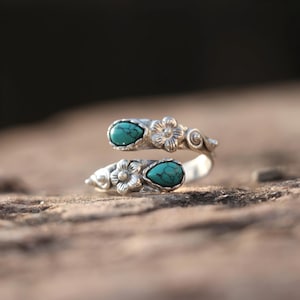 May include: A silver-toned ring with two teardrop-shaped turquoise stones. The ring features floral and swirl designs. The ring wraps around the finger, with the turquoise stones positioned on the top and bottom.