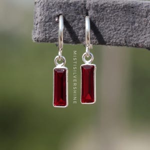 Può includere: Orecchini a cerchio in argento con pietre rettangolari rosso scuro. Gli orecchini pendono dai cerchi, su uno sfondo verde sfocato. Il testo "MISTISILVERSHINE" è visibile.