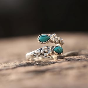 May include: A silver-toned ring with a floral design and two turquoise-colored stones. The ring wraps around the finger, with a flower and stone on each end. The ring is set on a textured surface.