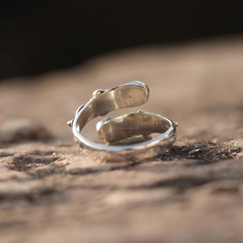 May include: A silver-colored adjustable ring with two oval-shaped settings. The ring has a textured band and is set against a blurred, natural background. The ring appears to be made of metal and is designed to be worn on a finger.