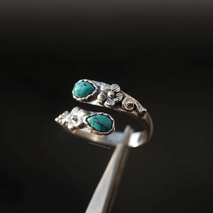 May include: A silver-toned ring with a floral design and two turquoise-colored teardrop-shaped stones. The ring has an open design, with the stones set on opposite ends. The ring is held by a silver-colored tool against a dark background.