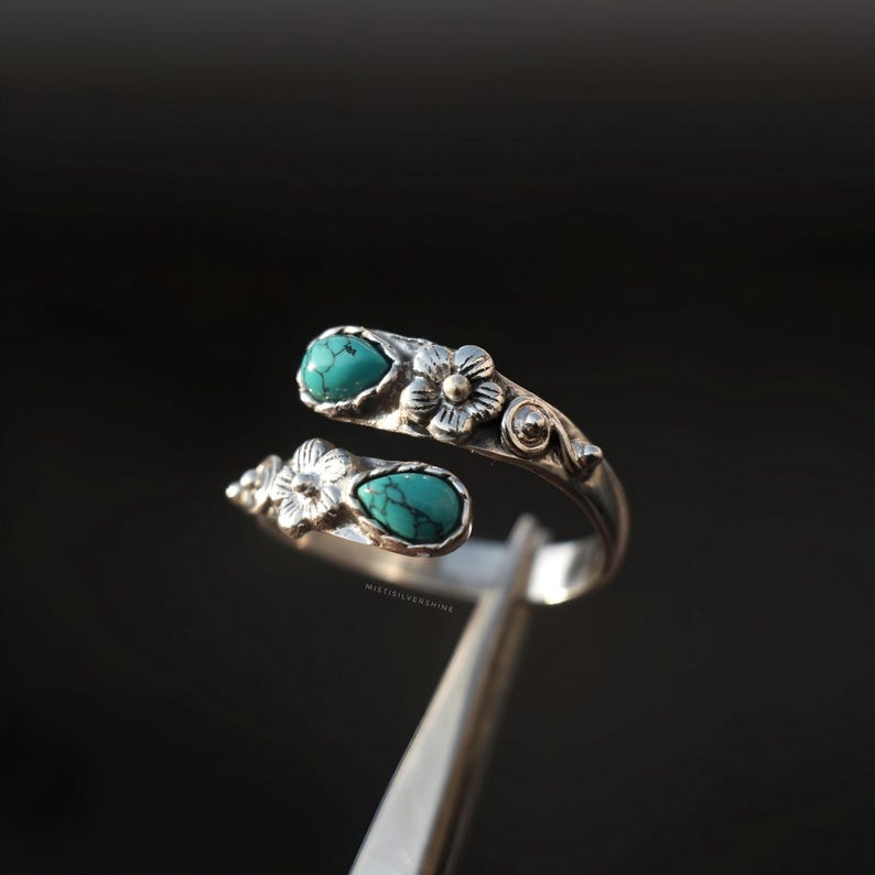 May include: A silver-toned ring with a floral design and two turquoise-colored teardrop-shaped stones. The ring has an open design, with the stones set on opposite ends. The ring is held by a silver-colored tool against a dark background.