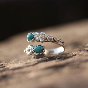 May include: A silver-toned ring with two turquoise-colored stones. The ring features floral and swirl designs. The stones are oval-shaped and set in a decorative setting. The ring is open and adjustable.