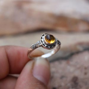 May include: A silver ring featuring a round, amber-coloured gemstone set in a circular frame with scalloped edges. The band displays intricate floral and leaf engravings on the sides. The text MISTISILVERSHINE is visible on the inner band.