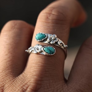 May include: A silver-toned ring with a floral design and two turquoise-colored stones. The ring wraps around the finger, with a flower and stone at each end. The ring is worn on a finger.