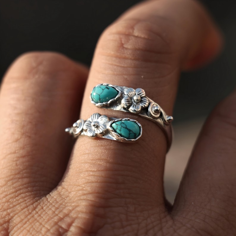 May include: A silver-toned ring with a floral design and two turquoise-colored stones. The ring wraps around the finger, with a flower and stone at each end. The ring is worn on a finger.
