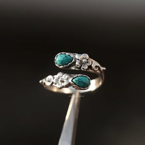May include: A silver-colored ring with two turquoise teardrop-shaped stones. The ring features floral and swirl designs. The ring is held by a silver tool against a dark background. The text "MISS SILVERSHINE" is visible.