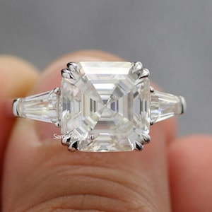 May include: A square-shaped diamond engagement ring with a white gold band. The center stone is a large, clear, square-cut diamond. Two smaller, baguette-cut diamonds are set on either side of the center stone.