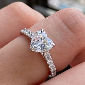 Può includere: Un anello d'argento con un diamante a forma di cuore incastonato al centro. La fascia è decorata con diamanti più piccoli.