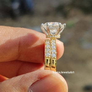 May include: A gold engagement ring set with a large, round, clear center stone and a halo of smaller stones. The band is also set with smaller stones. The ring is on a finger.