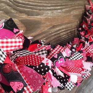 May include: A Valentine's Day wreath made of fabric strips in various patterns and colors. The wreath features red, pink, black, and white fabrics with polka dots, hearts, and gingham patterns. The wreath is displayed against a rustic wooden background.