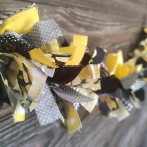 May include: A handmade fabric garland featuring yellow, black, and gray fabric strips. The garland includes solid yellow, polka dot, and patterned fabric with a lemon theme. The decorative item is displayed against a wooden background.