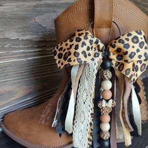 May include: A brown western-style boot adorned with a decorative accessory featuring a leopard print bow, layered ribbons in cream, black, and tan, lace trim, and a string of beads including a leopard print mouse-shaped charm with a bow.