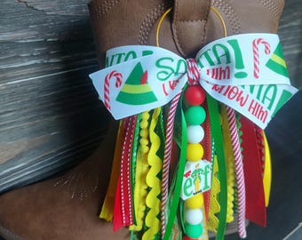 Puede incluir: Una bota vaquera marrón adornada con un lazo decorativo con el texto SANTA! I KNOW HIM, bastones de caramelo y gorros de elfo. El lazo incluye cintas colgantes en rojo, amarillo y verde, junto con una cadena de cuentas y un dije de elfo.