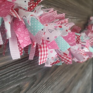 May include: A handmade fabric garland featuring a variety of pink, red, and white patterned fabrics. The garland includes fabrics with heart, polka dot, and gingham patterns, and a few pieces of light blue fabric. The garland is likely for Valentine's Day or a similar celebration.