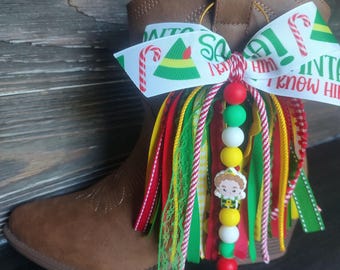 Puede incluir: Una bota vaquera marrón adornada con un adorno colgante festivo que presenta una cinta blanca estampada con bastones de caramelo, gorros de elfo y el texto 'SANTA! I KNOW HIM!'. El adorno incluye cintas coloridas y cuentas con un elfo.