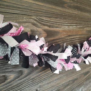 May include: A handmade fabric garland featuring alternating sections of pink, black, and gray fabric strips. Some strips have white polka dots. The garland is displayed against a rustic wooden background.