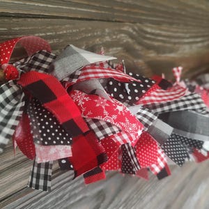 May include: A decorative garland made of numerous tied fabric strips in red, black, and white. The fabric patterns include buffalo plaid, small gingham checks, white polka dots on black, and red fabric featuring white snowflake designs.