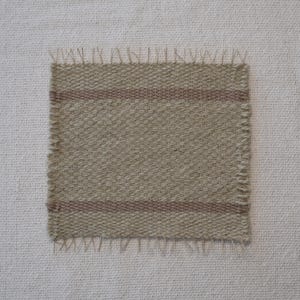 May include: A square, woven fabric swatch with a beige and brown color scheme. The fabric has a textured appearance with two horizontal brown stripes. The edges are fringed, and the swatch is set against a white background.