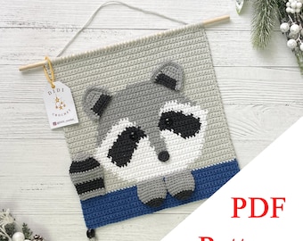 May include: Crocheted wall hanging featuring a raccoon design. The raccoon has gray, white, and black details, with a blue border at the bottom. The hanging is attached to a wooden dowel with a string for display. The text "PDF Pattern" is visible.