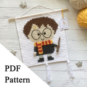 May include: A white crochet wall hanging featuring a cartoon character with brown hair, glasses, and a red and yellow scarf. The character is holding a wand. The wall hanging is suspended from a wooden dowel and has white tassels.