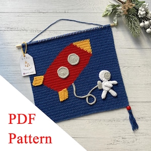 May include: A blue crochet wall hanging featuring a red rocket with yellow accents and two gray circular windows. A white astronaut is connected to the rocket by a gray cord. The text "PDF Pattern" is visible.