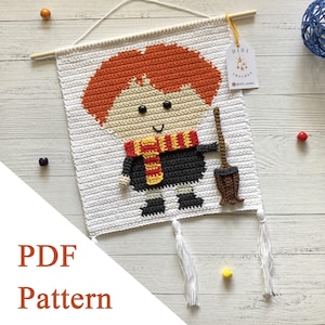 May include: Crocheted wall hanging featuring a cartoon character with red hair, a scarf, and a broom. The character is on a white background with a wooden dowel at the top. The text "PDF Pattern" is visible. The wall hanging has white tassels.