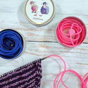 Peut inclure: Trois petites boîtes rondes en métal présentent différents contenus. L'une contient des élastiques bleus, une autre des élastiques roses, et la troisième affiche une illustration de mouton avec le texte "Zazouch Créations". Un projet tricoté en violet et blanc est en cours.