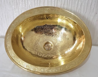 May include: A gold oval-shaped bathroom sink with an ornate engraved design around the rim. The sink has a hammered finish and a drain in the center.
