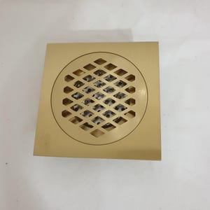 May include: A square-shaped gold-colored metal floor drain cover featuring a circular center with a diamond-patterned lattice grate. The design includes a smooth flat border surrounding the central grid, which reveals a perforated metal layer underneath.