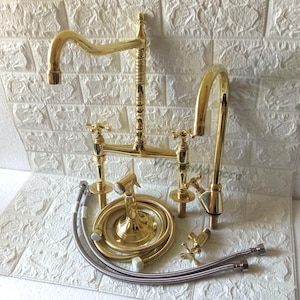 May include: A complete gold-coloured kitchen tap set. The set includes a bridge tap with cross handles, a side sprayer, and flexible supply lines. The tap has a curved spout and a decorative design.