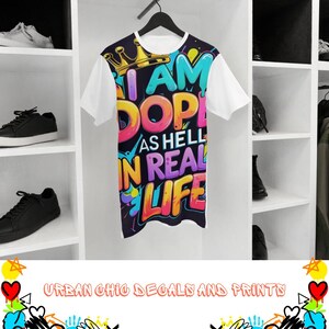 May include: White short-sleeve t-shirt with a colorful graphic that reads "I AM DOPE AS HELL IN REAL LIFE." The design includes a gold crown and vibrant lettering in blue, pink, yellow, and purple. The shirt is hanging on a black hanger.
