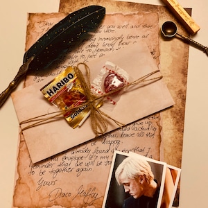May include: A vintage-style letter set with handwritten text, a feather quill, and a small photo. The set includes a sealed envelope tied with twine, a bag of gummy candies, and a heart-shaped candy. A wax seal and spoon are also present.