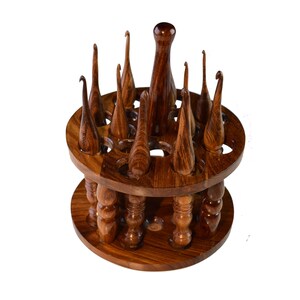 May include: A wooden stand with a rotating base holds a collection of wooden crochet hooks. The hooks are arranged in a circular pattern around the stand, with the tips of the hooks pointing upwards.