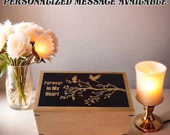 May include: A wooden box with a black lid featuring a gold silhouette of a tree branch with birds and the words "Forever In My Heart". The box is on a white surface with a lit candle and white flowers in a vase.