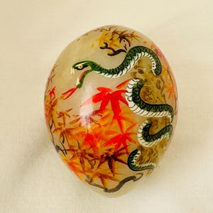 Puede incluir: Una piedra blanca en forma de huevo con un diseño pintado a mano de una serpiente verde que se enrosca en una rama de hojas de arce rojas y amarillas.