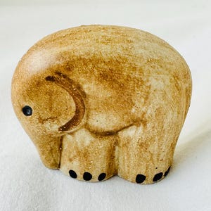 May include: A small, ceramic elephant figurine in shades of tan and brown. The elephant has a rounded body, a curved trunk, and small black dots representing its feet. The figurine has a textured surface and a simple, charming design.