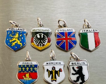 May include: Seven enamel and silver charm pendants featuring shields with various national and city emblems. The shields are decorated with colorful enamel and feature images such as a lion, eagle, bear, and a ship. The charms are labeled with the names "Holland", "Deutschland", "England", "Italien", "Paris", "München", and "Berlin".