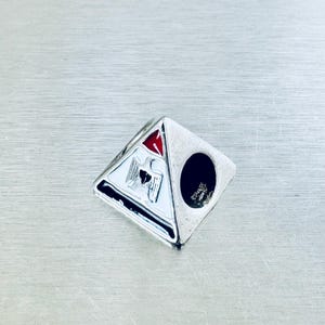 May include: Silver-toned pyramid-shaped ring with a black circle and a white triangle with a red triangle and an eagle emblem. The ring has a detailed design and is set against a silver background.