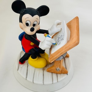 Puede incluir: Una figura de cerámica de Mickey Mouse sentado en una silla azul frente a una mesa de dibujo de madera. Mickey tiene orejas negras, cara blanca, pantalones rojos con botones amarillos y zapatos amarillos. Sostiene un lápiz azul sobre papel.