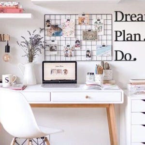 May include: A white desk with a white chair, a laptop, a lamp, a vase of lavender, and a grid wall with photos and the words "Dream. Plan. Do." on the wall.