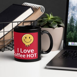Può includere: Tazza da caffè nera con un pannello rosso con una faccina gialla sorridente e la scritta "I Love Coffee HOT" in bianco. La tazza è su una superficie bianca con un laptop, libri e una pianta sullo sfondo.