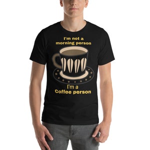 Puede incluir: Camiseta negra con un diseño gráfico con temática de café. El diseño presenta una taza de café y un platillo con el texto "I'm not a morning person, I'm a Coffee person" en amarillo. La camiseta la lleva una persona.