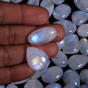 May include: A collection of iridescent white moonstone cabochons in various teardrop and oval shapes. The stones have a milky white color with a blue and rainbow sheen.
