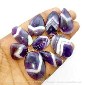 K&ouml;nnte beinhalten: Eine Sammlung von zehn violetten und wei&szlig;en Amethyst-Cabochons mit Chevron-Mustern. Die Cabochons sind in verschiedenen Formen und Gr&ouml;&szlig;en und werden auf einer Hand pr&auml;sentiert.