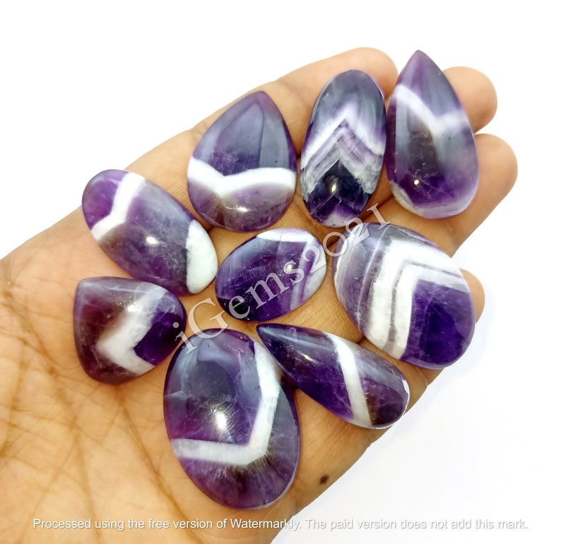 K&ouml;nnte beinhalten: Eine Sammlung von zehn violetten und wei&szlig;en Amethyst-Cabochons mit Chevron-Mustern. Die Cabochons sind in verschiedenen Formen und Gr&ouml;&szlig;en und werden auf einer Hand pr&auml;sentiert.