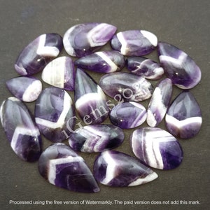 K&ouml;nnte beinhalten: Eine Sammlung von 17 Amethyst-Cabochons mit wei&szlig;en Chevron-Mustern. Die Cabochons sind tropfenf&ouml;rmig und haben eine polierte Oberfl&auml;che.
