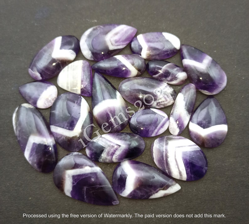 K&ouml;nnte beinhalten: Eine Sammlung von 17 Amethyst-Cabochons mit wei&szlig;en Chevron-Mustern. Die Cabochons sind tropfenf&ouml;rmig und haben eine polierte Oberfl&auml;che.