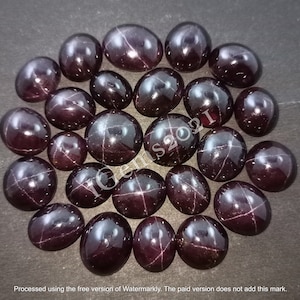 May include: A collection of 20 dark red star-shaped gemstones, also known as star garnets, arranged in a circle on a dark brown surface. The gemstones are oval-shaped and have a star-like pattern in the centre.